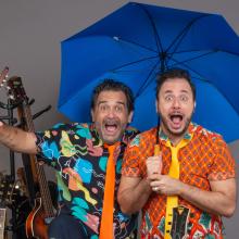 foto de 2 artistas homens com suas bocas abertas, 1 está segurando um guarda-chuva azul aberto e atrás do lado esquerdo tem algumas guitarras